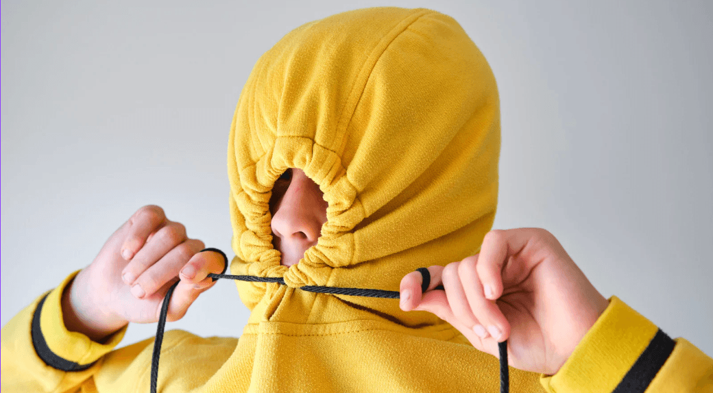 Kid in yellow hoodie with the hood cinched closed tight pulling the strings with both hands, as if hiding from social media shame.